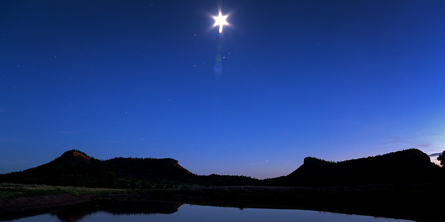 White star-shaped ball of light in dark blue sky over silhouette of two black buttes and reflecting pond in the foreground