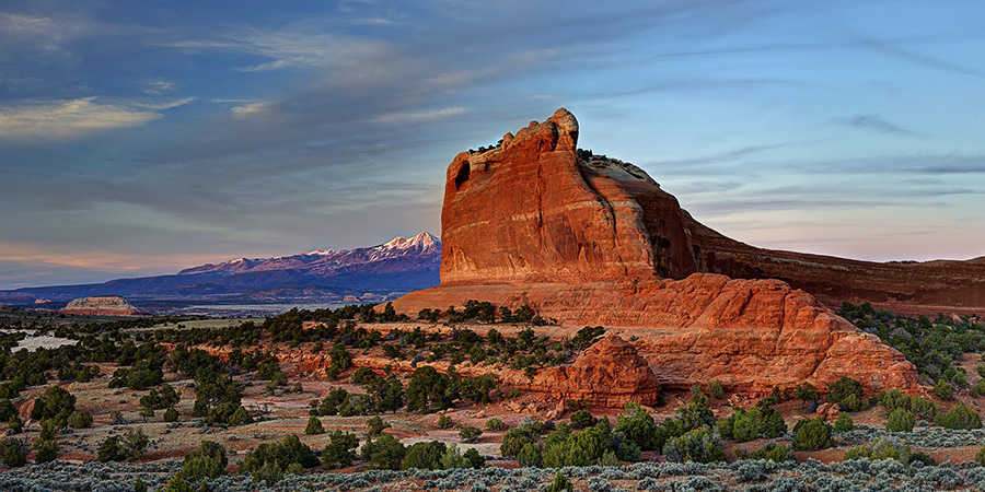 Wind Whistle Rock, a red rock butte with pinyon and juniper trees dotting the base and purple snow capped mountains in the distance