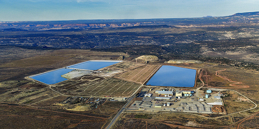Bright blue ponds of the White Mesa uranium mill on flat brown expanse of black mesa with forested hills in the distance