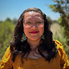 A person with long dark hair and glasses, wearing a yellow shirt and turquoise earrings, stands outdoors with greenery in the background.