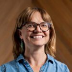 A woman with shoulder-length light brown hair and glasses smiles, wearing a blue denim shirt and green earrings, with a blurred brown background.