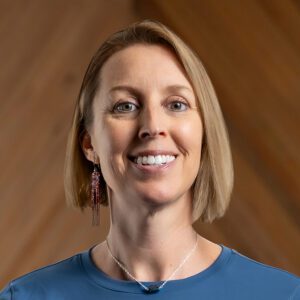A woman with straight blonde hair, wearing a blue shirt, long beaded earring, and a necklace, smiles in front of a wooden background.
