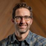 Michael Toll, a man with curly brown hair, glasses, and a beard, wearing a plaid shirt, is smiling in front of a blurred wooden background.