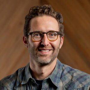 Michael Toll, a man with curly brown hair, glasses, and a beard, wearing a plaid shirt, is smiling in front of a blurred wooden background.