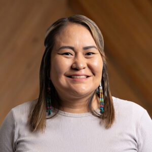 A woman with straight, shoulder-length hair and beaded earrings smiles at the camera, wearing a light-colored ribbed top against a wooden background.