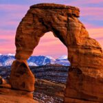 Delicate Arch, a large red rock arch in Arches National Park with dramatic pink and purple sky and snow-capped mountains in the background