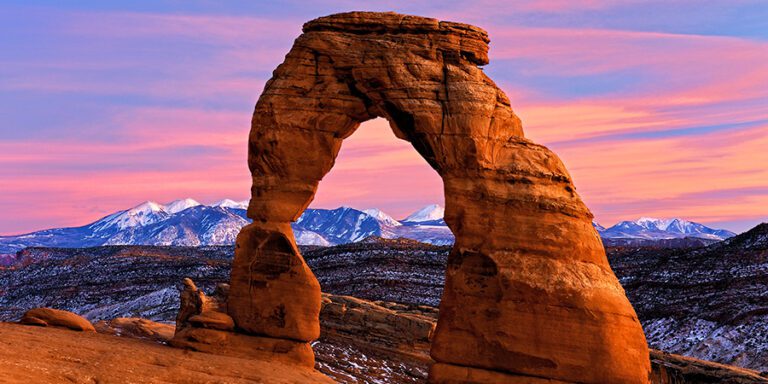 Delicate Arch, a large red rock arch in Arches National Park with dramatic pink and purple sky and snow-capped mountains in the background