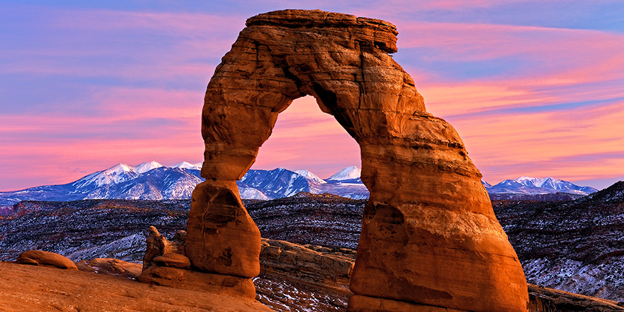 Delicate Arch, a large red rock arch in Arches National Park with dramatic pink and purple sky and snow-capped mountains in the background