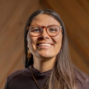 Kaya McAlister, a woman with long brown hair, glasses, and dangling earrings, smiles at the camera against a wooden background.