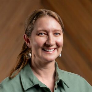 A woman with light brown hair, wearing a green collared shirt and dangling earrings, smiles at the camera against a wooden background.