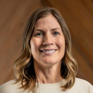 Melanie Seus, a woman with shoulder-length blonde hair, wearing a light-colored top, smiles at the camera in front of a wooden background.