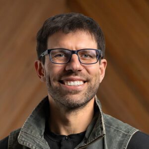 A man with short brown hair, glasses, and a beard smiles at the camera. He is wearing a gray jacket over a black shirt, with a wooden background behind him.