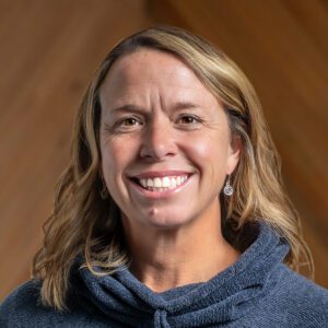 A woman with wavy blonde hair, wearing a blue sweater and drop earrings, smiles in front of a wooden background.