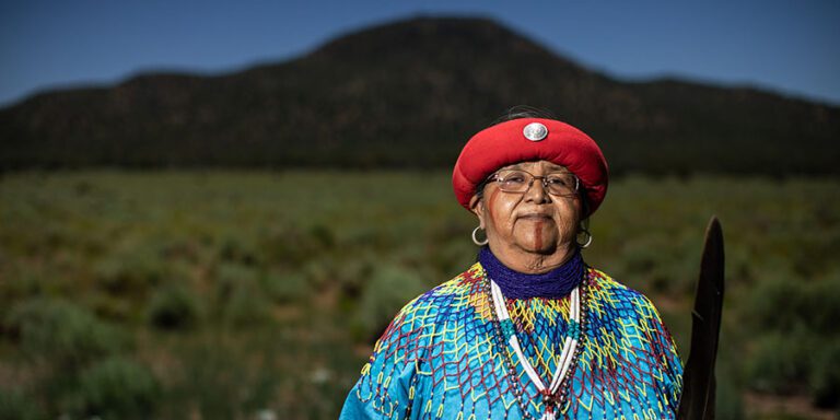 Dianna Sue White Dove Uqualla, a Native American woman in traditional clothing and a red headcovering stands in a grassy field with a mountain, Red Butte, in the background, holding a feather.