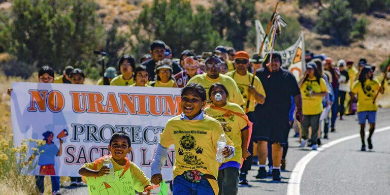 A group of people march along a road holding banners and signs, including one that reads "No Uranium; Protect Sacred Land." Children lead the march, and many participants wear yellow shirts.