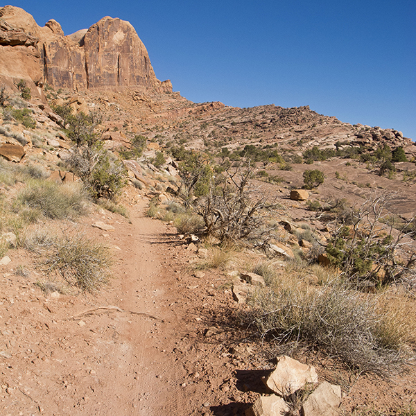 A desert trail on red dirt skirts below sandstone cliffs