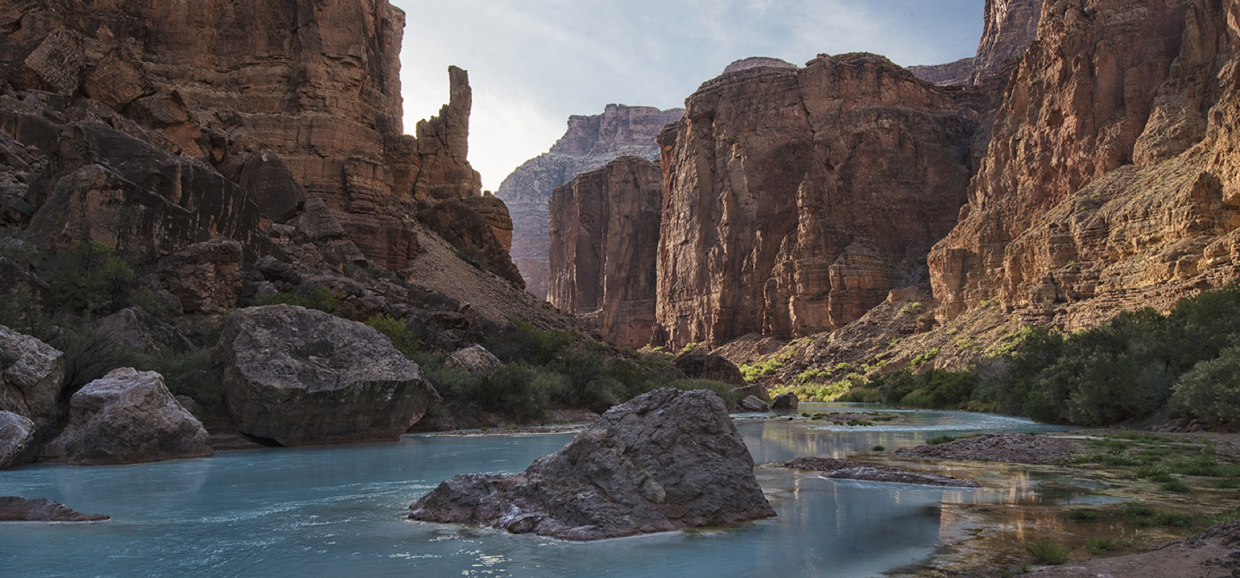 Milky turquoise river flows around large rock with high reddish cliffs on both sides, the Little Colorado River near the Grand Canyon