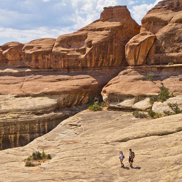 Two hikers walk on a ramp of slickrock beneath sandstone towers.