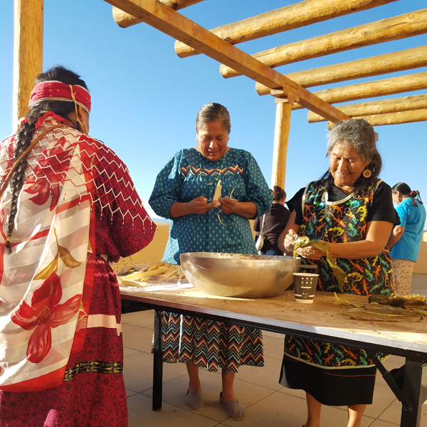 Three women prepare traditional food underneath a wooden shade structure.
