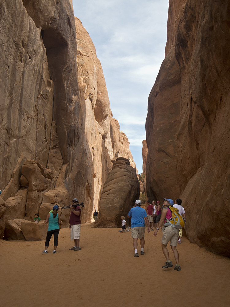 Hikers stand between two narrow sandstone walls, with a large boulder in the middle.