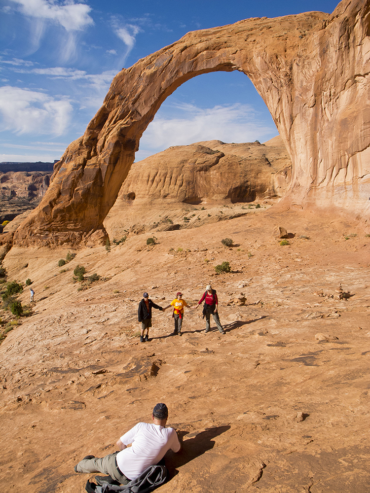 A man in a white shirt lays on the slickrock to take photos of his three hiking companions under the arch