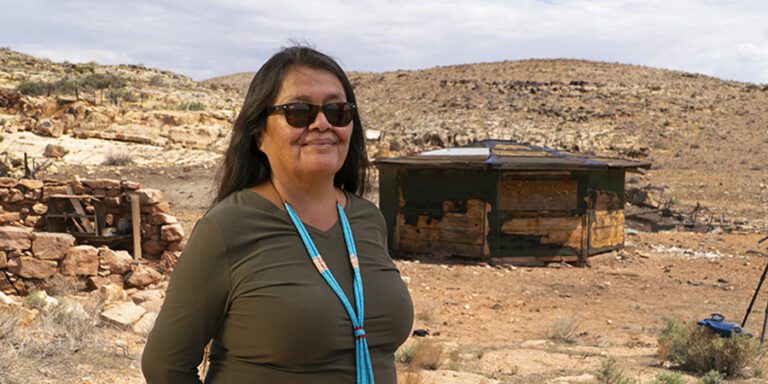 Woman with long dark hair and turquoise necklace smiling and wearing sunglasses.