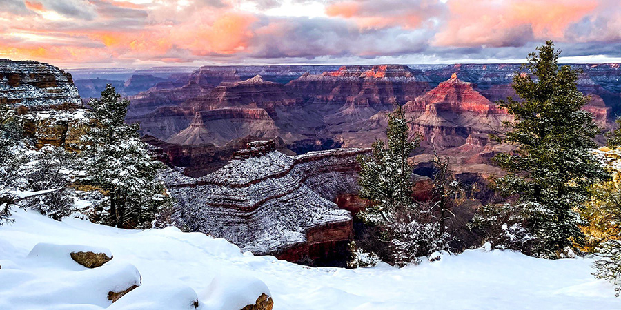 The last reddish rays of sunlight illuminate buttes and cliffs within an immense canyon topped with snow.