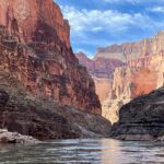 Clear calm green Colorado River with step red, pink, brown, and orange Grand Canyon cliffs on both sides.