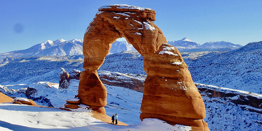Delicate Arch, large red rock arch with two people underneath and snowy mountains in the background, Arches National Park,