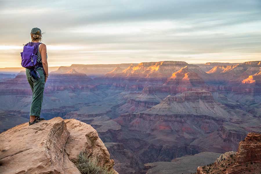 Woman in a purple backpack stands looking out at the purple-colored rocks of the grand canyon at sunrise