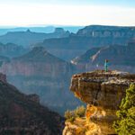 Woman in bright clothes stands on a rock outcropping looking out at the Grand Canyon whose rock layers fade to blue in the distance
