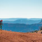 Backpacker in bright clothing walks along a rocky red plateau with the blue layers of the Grand Canyon in the background.