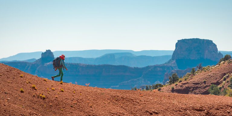 Backpacker in bright clothing walks along a rocky red plateau with the blue layers of the Grand Canyon in the background.
