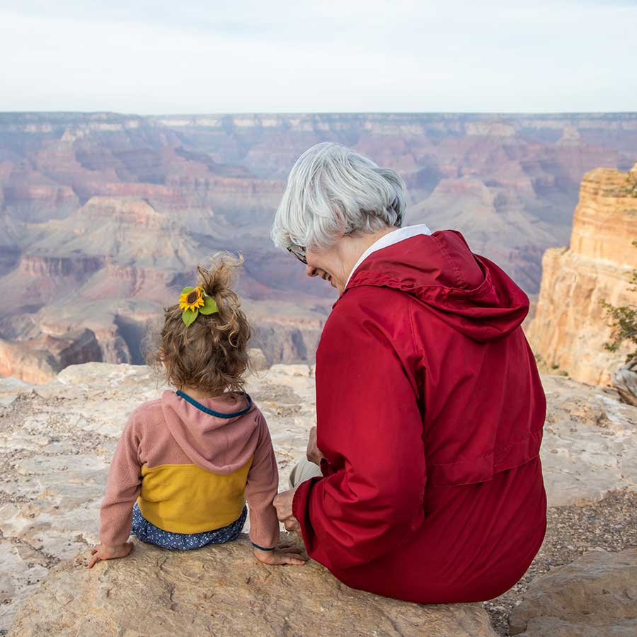 A grandma in a red jacket and a little girl with a ponytail sit together on a rock and admire the Grand Canyon