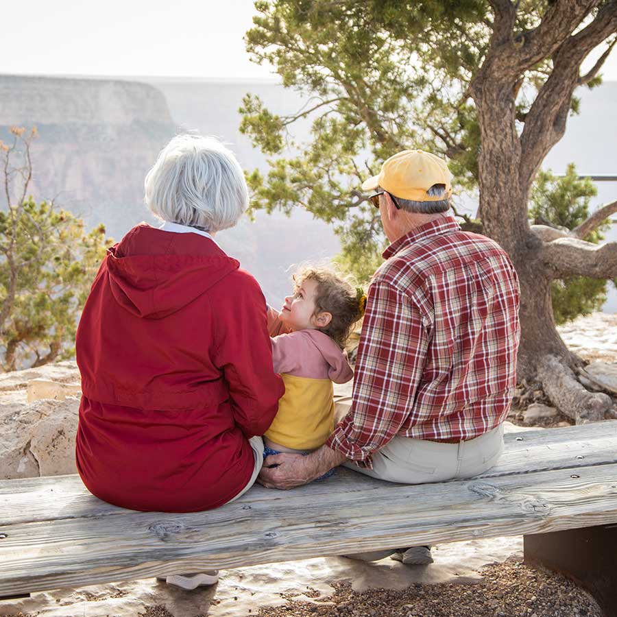 A granddaughter sits between her two grandparents on a bench overlooking the rim of the Grand Canyon