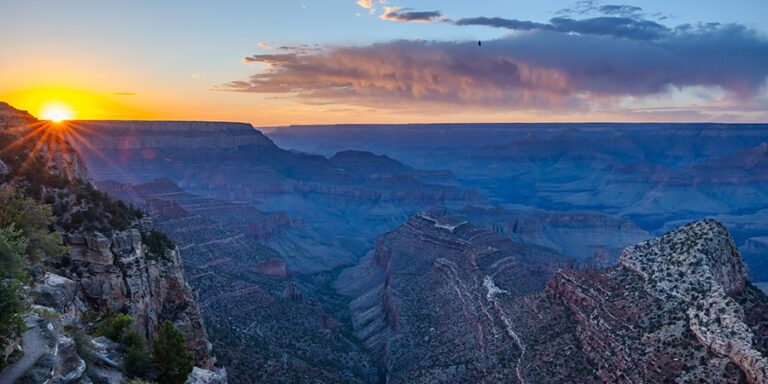 A starburst sun sets behind the cliffs at Grandview Point, casting an orange and blue glow across the desert landscape.