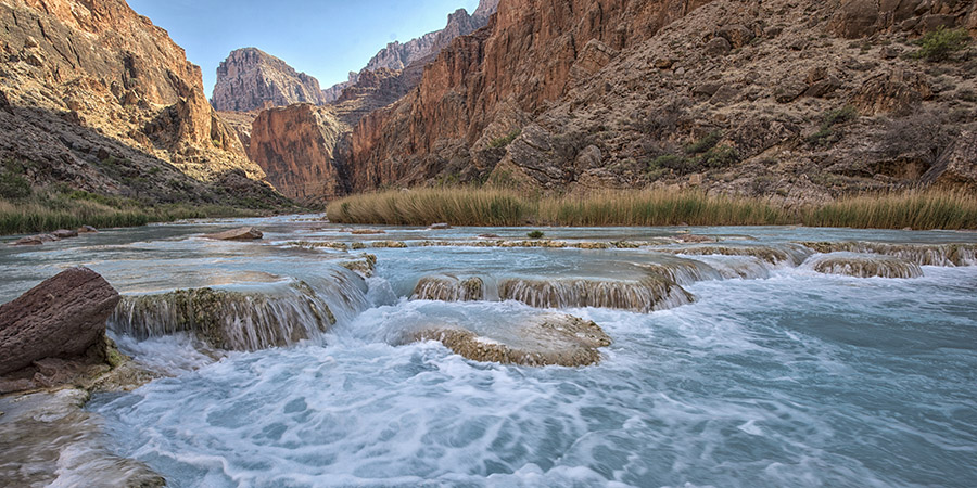 Travertine dams with turquoise blue warm carbonate water of the Little Colorado River flowing over them and foaming white, with canyon walls rising