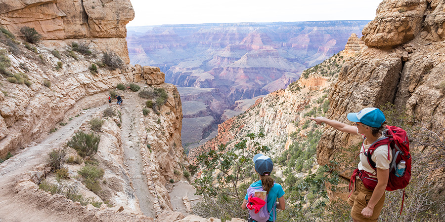 Two hikers with backpacks stand on a rocky trail overlooking the Grand Canyon, while one points towards the canyon in the distance—an inspiring scene perfect for illustrating the wonders of the Grand Canyon for teachers and students alike.