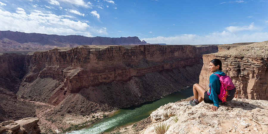 A person wearing a pink backpack sits on a rocky ledge overlooking the Baaj Nwaavjo Attack site, where a river winds through a deep canyon under a partly cloudy sky.