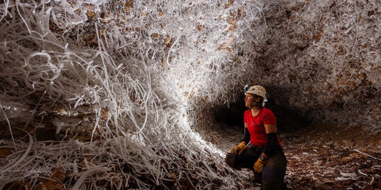 Woman in red shirt and helmet looks at white glass-like formations in a cave.