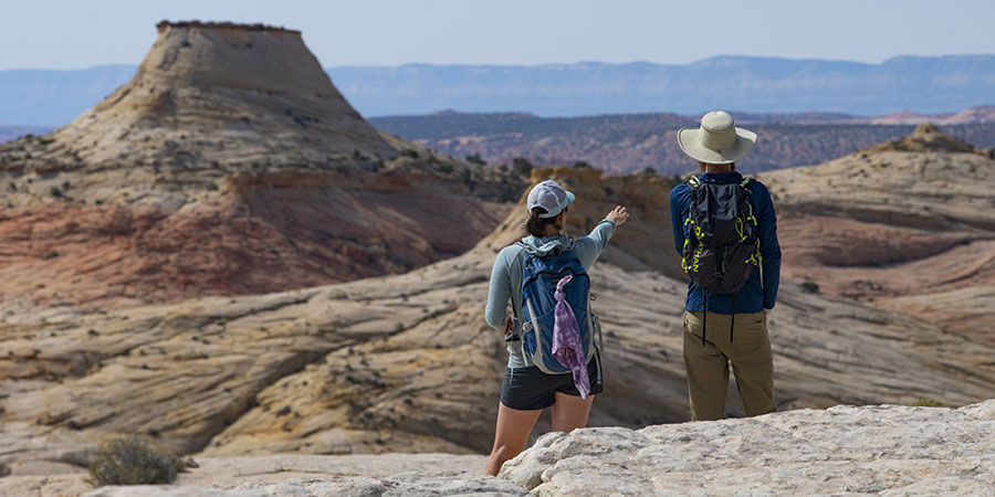 A male and female hiker on orange and white sandstone point toward a plateau in the distance.