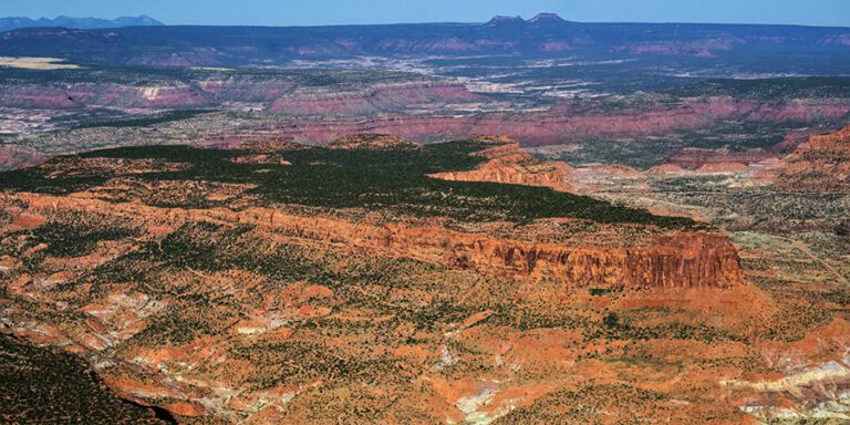 Aerial view of a vast landscape near the Daneros uranium mine, showcasing red rock plateaus, mesas, and scattered vegetation under a clear blue sky with the Bears Ears buttes in the background.