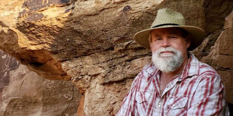 A man with a gray beard wearing a straw hat and plaid shirt sits in front of a rocky, sunlit cliff.