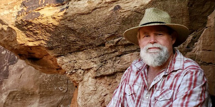 A man with a gray beard wearing a straw hat and plaid shirt sits in front of a rocky, sunlit cliff.