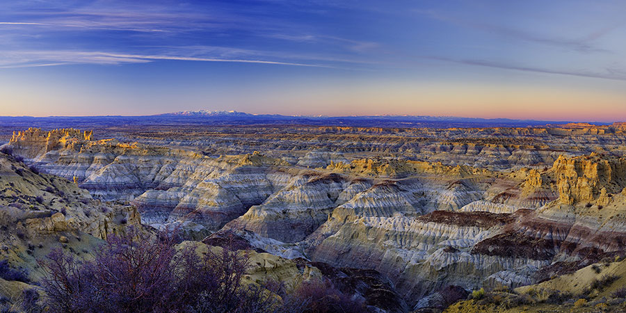 Wide view of a rugged badlands landscape with layered rock formations and distant mountains under a clear sky at sunrise or sunset.