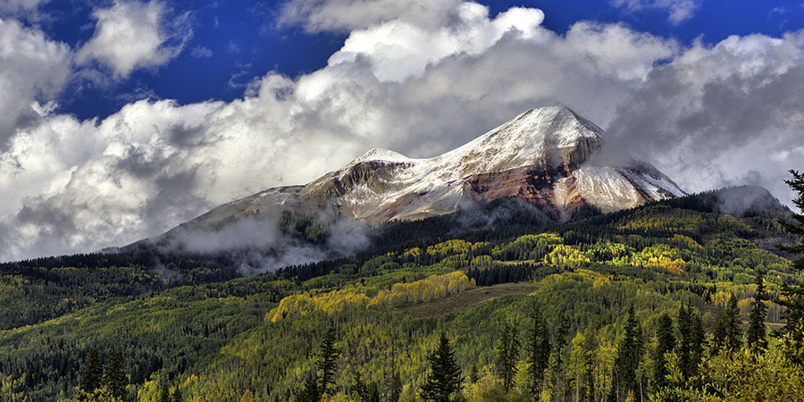 Snow-capped mountain peak under partly cloudy sky, with dense forests and patches of yellow-green trees covering the lower slopes.