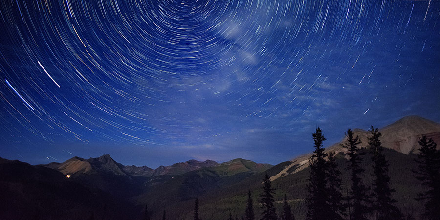 Long-exposure photo of a night sky with circular star trails above mountains and forested hills.