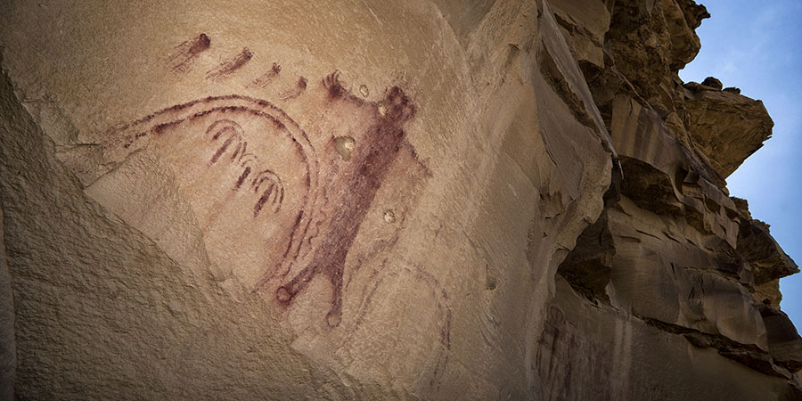 Red-brown pictographs depicting a human-like figure and arc shapes on a large sandstone cliff face under a clear sky.