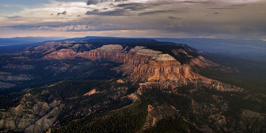 Aerial view of a rugged, forested mesa with layered red and white rock formations under a cloudy sky.