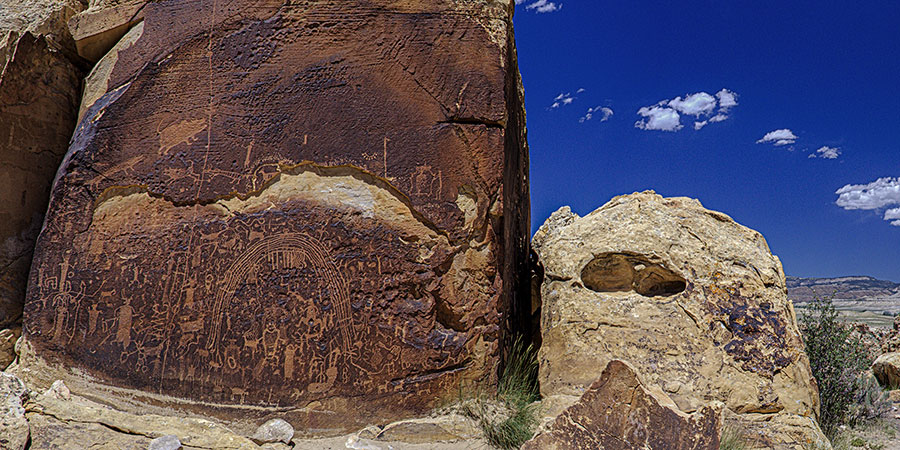 Petroglyphs are carved into a large rock face under a clear blue sky, with another light-colored boulder nearby.